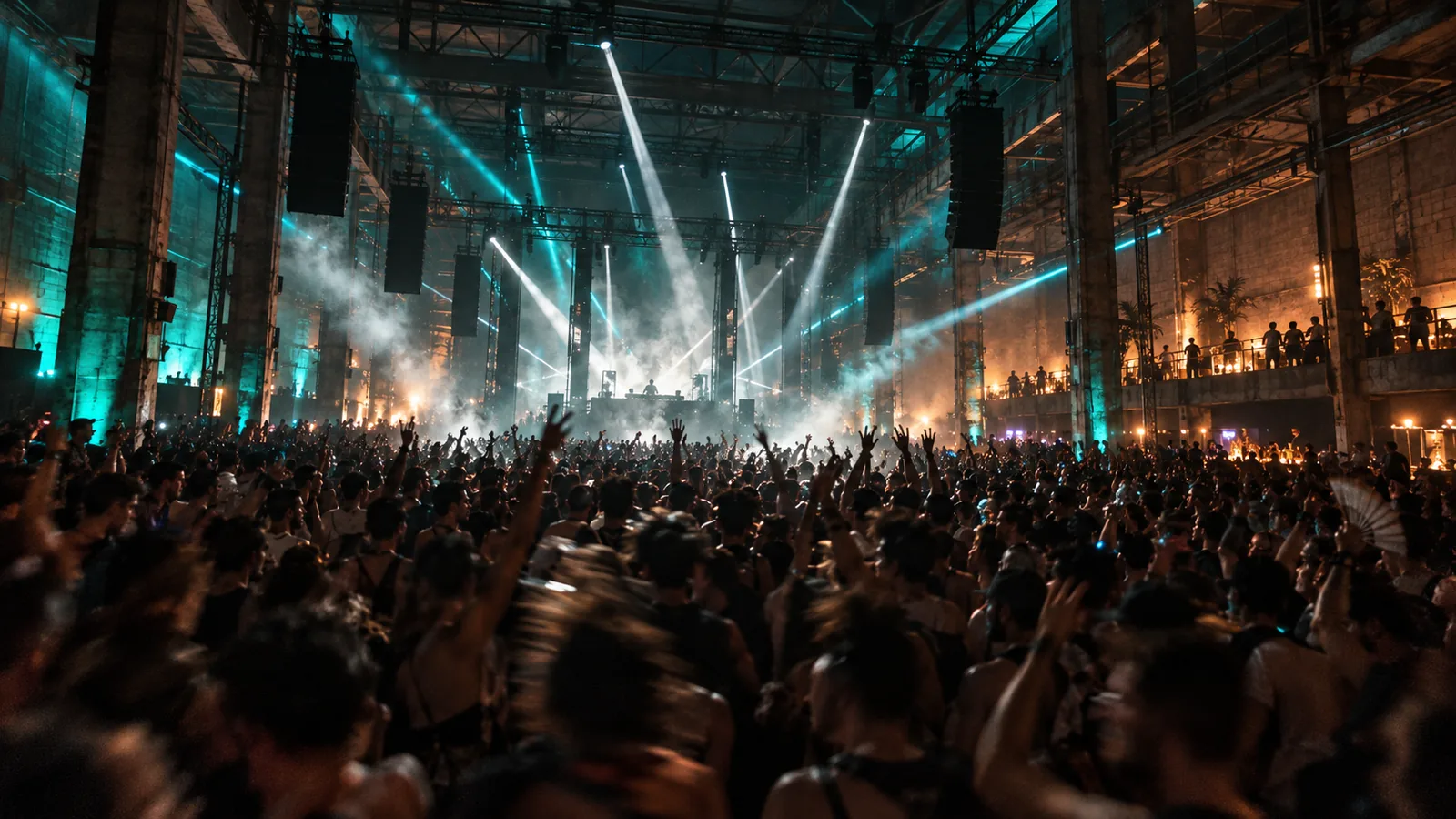 A warehouse room packed to the stage, hands up under steel trusses and white and teal light beams.
